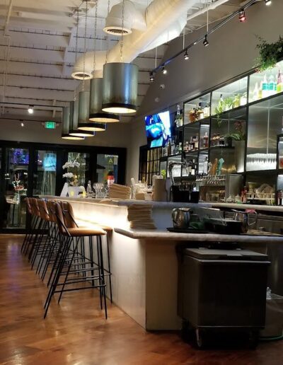 A modern bar with high wooden chairs and hanging cylindrical lights. The bar counter is stocked with bottles and glassware. The atmosphere is bright with wooden flooring and an industrial ceiling.