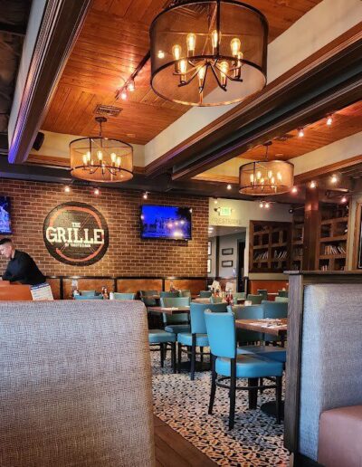 A restaurant interior with wooden ceiling, brick walls, pendant lighting, blue chairs, and booth seating. The GRILLE sign and TV screens are visible on the wall. A person is standing near the counter.