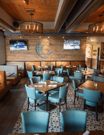 The image shows the interior of a restaurant with blue chairs, wooden tables, and brick walls. There are two TV screens on the wall, and large windows provide natural light.