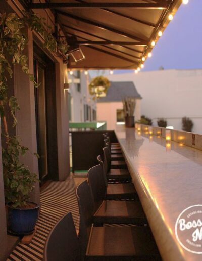 A long, marble counter with barstools on an outdoor patio, adorned with string lights at dusk. A "Bossa Nova" logo is visible in the bottom right corner. Plants decorate the left side.