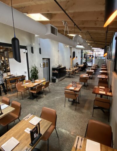 A modern restaurant interior with wooden tables, brown leather chairs, and industrial-style lighting. The restaurant appears empty, with neatly set tables and a visible kitchen area in the background.