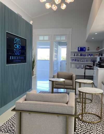 Modern waiting room with cushioned chairs, tall plants, wall-mounted TV displaying "Executive Settings," and well-lit shelves with various items.