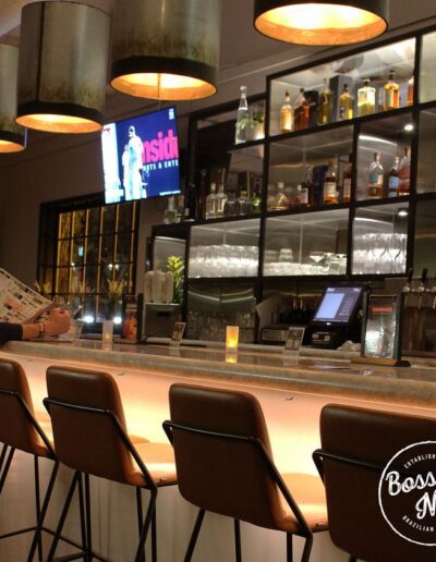 A modern bar with high chairs, shelves with glasses and liquor bottles, a TV screen, a person reading at the bar, and a lit up "Bossa Nova" sign in the corner.