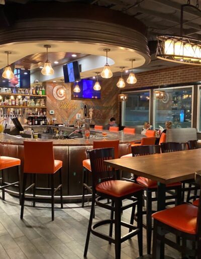 A modern bar with an oval-shaped counter and orange chairs. Customers are seated at the counter, and the bar is stocked with various bottles. The background shows a lit sign and a flat-screen TV.