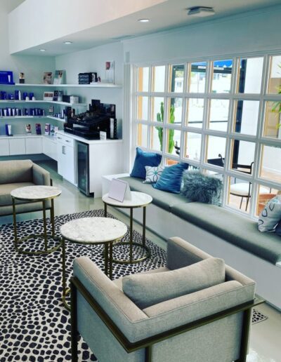 A modern waiting area with gray chairs, a cushioned window seat, small round tables, and a patterned rug. Bookshelves and a refreshment counter are visible in the background. Bright natural light.