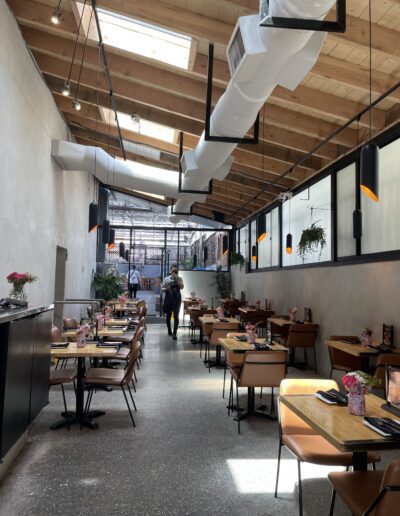 A modern, sunlit restaurant with wooden ceiling beams, ductwork, and tables set with flowers. A few people are walking.