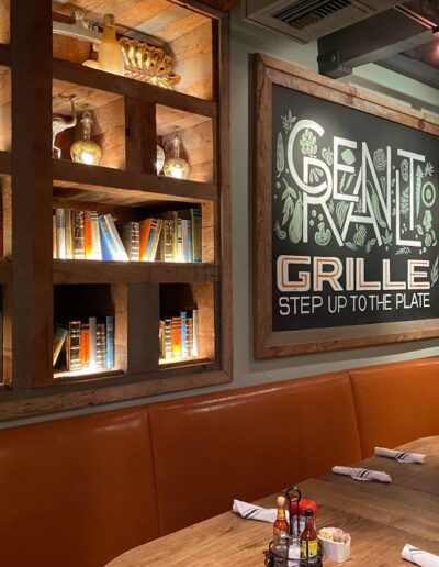 A restaurant interior with a lit bookshelf display, framed "Senate Grille" sign, and a wooden table set for four with napkins, cutlery, and condiments. Tan cushioned seating lines the wall.