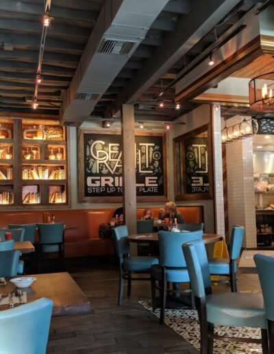 Interior of a restaurant with wooden tables, blue cushioned chairs, exposed ceiling beams, and a well-lit kitchen area. Shelves with decorative items and a sign reading "Grille" adorn the back wall.