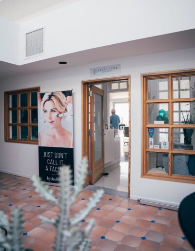 Image of a spa entrance with a signboard displaying a woman's face and text reading "Just Don't Call It a Facial." A person in blue scrubs stands inside the spa, near treatment rooms.