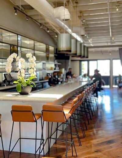 A modern restaurant interior featuring a long bar with high chairs, pendant lighting, and a sleek design aesthetic. The space has polished wood flooring and an arrangement of orchids on the bar.