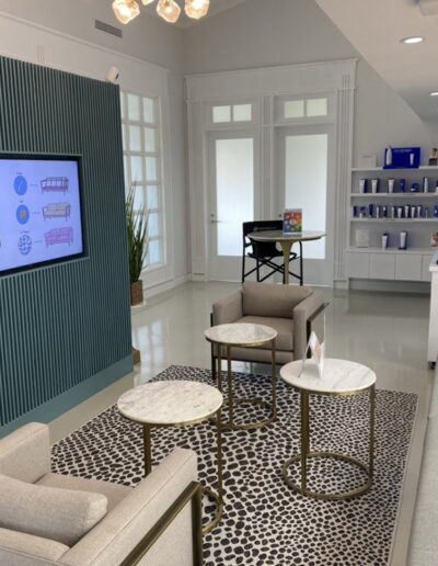 Modern waiting area with a wall-mounted TV, comfortable seating, and small tables. A shelf in the background holds various products. Large windows allow natural light to fill the room.