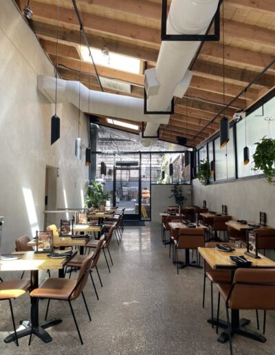 A modern, empty restaurant with wooden tables and brown chairs. Exposed ductwork is visible on the ceiling. Sunlight filters through skylights and large side windows, illuminating the space.